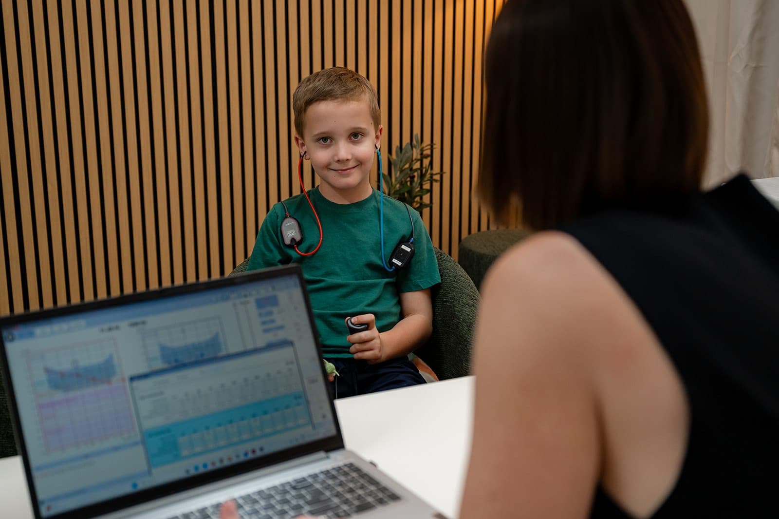 A child wearing insert earphones and holding the response button during a hearing test, with the audiogram on the laptop