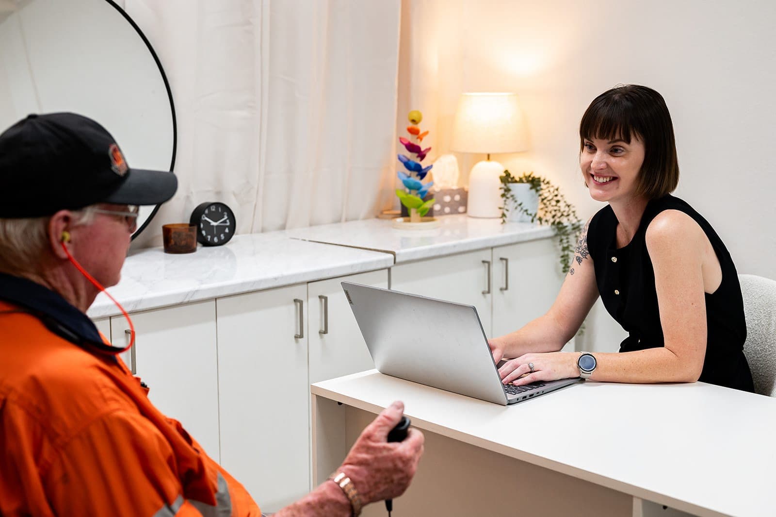A worker in orange hi-vis wearing insert earphones during a workplace hearing test, with Dr Alison Collins at the laptop